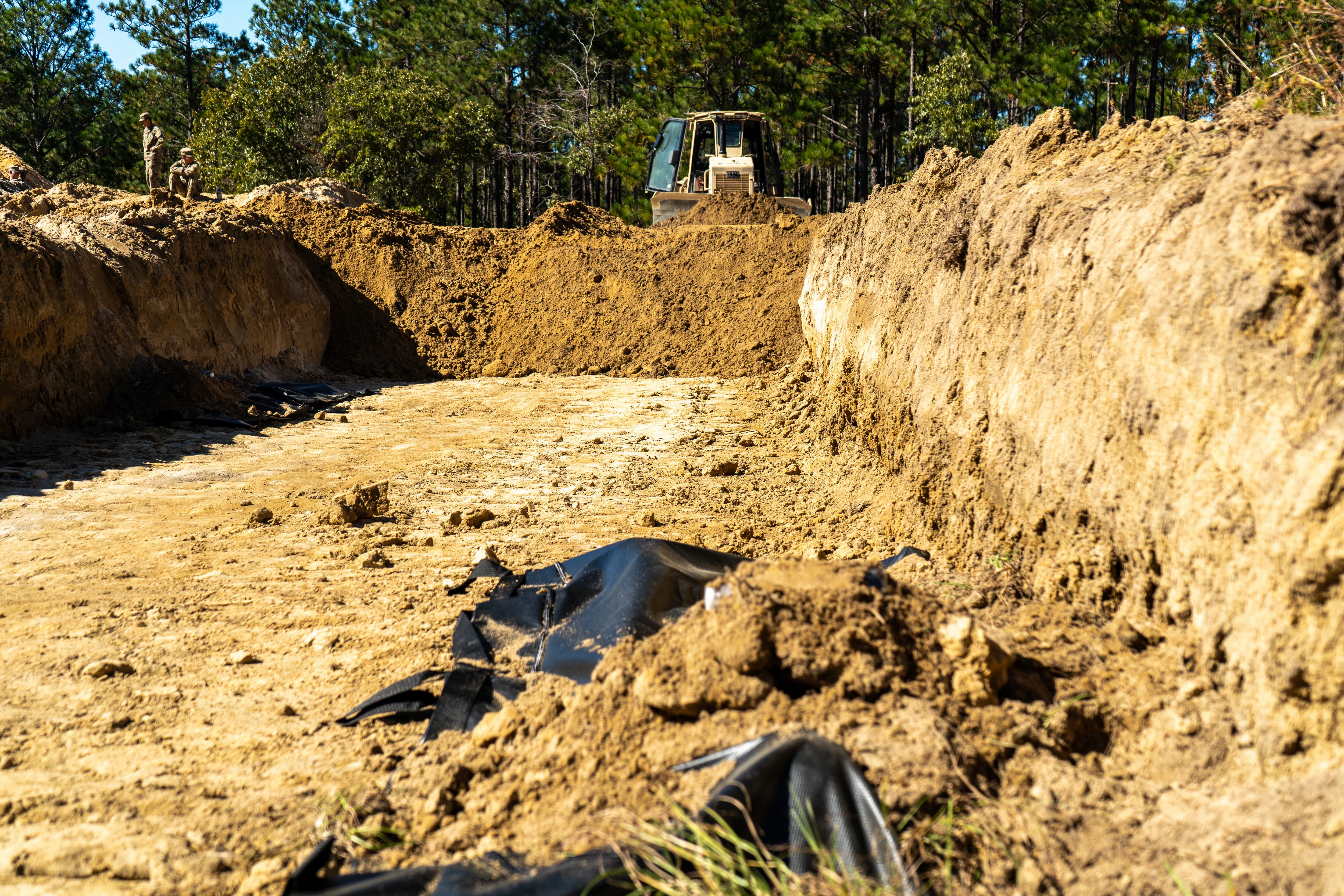 A U.S. Army D6 bulldozer, operated by horizontal construction engineers from the 20th Engineer Brigade, backfills a simulated temporary interment trench during Phase II of Operation Dragon Covenant near the St. Mere Eglise Drop Zone, Oct. 21, 2026. The test-of-concept exercise, executed by the 3rd Corps Sustainment Command, evaluated the structural impact of heavy earthmoving equipment on remains by placing simulated skeletal models and sealed water containers inside burial bags. Operators practiced casting dirt inward from a 15-foot-wide trench to verify that the gravity fall of displaced soil preserves remains intact for future forensic identification and repatriation, while refining spatial standards for future Army fatality management doctrine. (U.S. Army photo by Capt. Thomas McCarty)